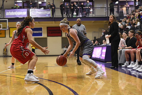 Photo of female Chatham University basketball player dribbles the ball against an opponent in a full arena. 