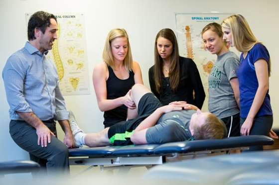 Photo of an instructor and four female physical therapy students at Chatham University surrounding a patient on a table as one student adjusts his leg. 
