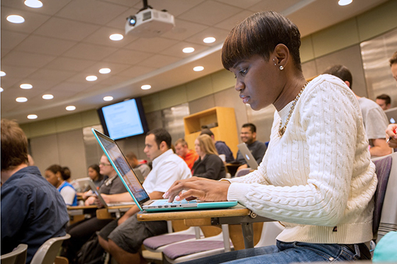 Photo of a Chatham University student sitting in full lecture hall at Chatham Eastside, taking notes on her laptop. 