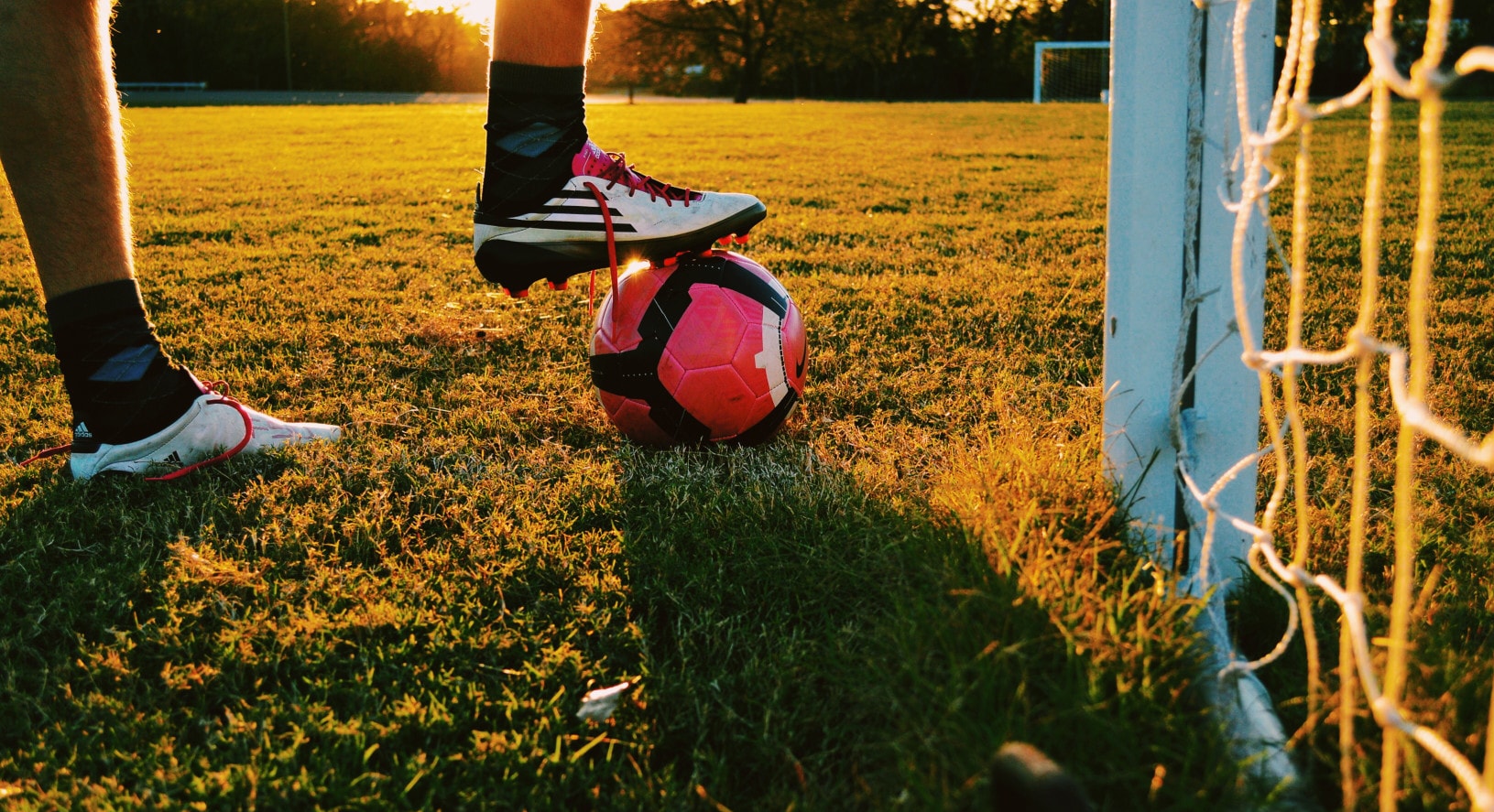 Photo of a soccer player wearing cleats, resting a foot on a soccer ball on a field next to a goal. The lighting in the photo suggests sunset.