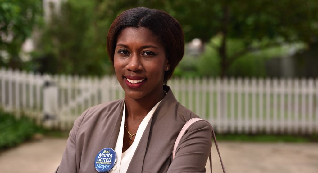 Photo of Chatham University's Marita Garrett, Mayor of the Borough of Wilkinsburg, posing in a brown blazer with a campaign button and arms crossed outside. 
