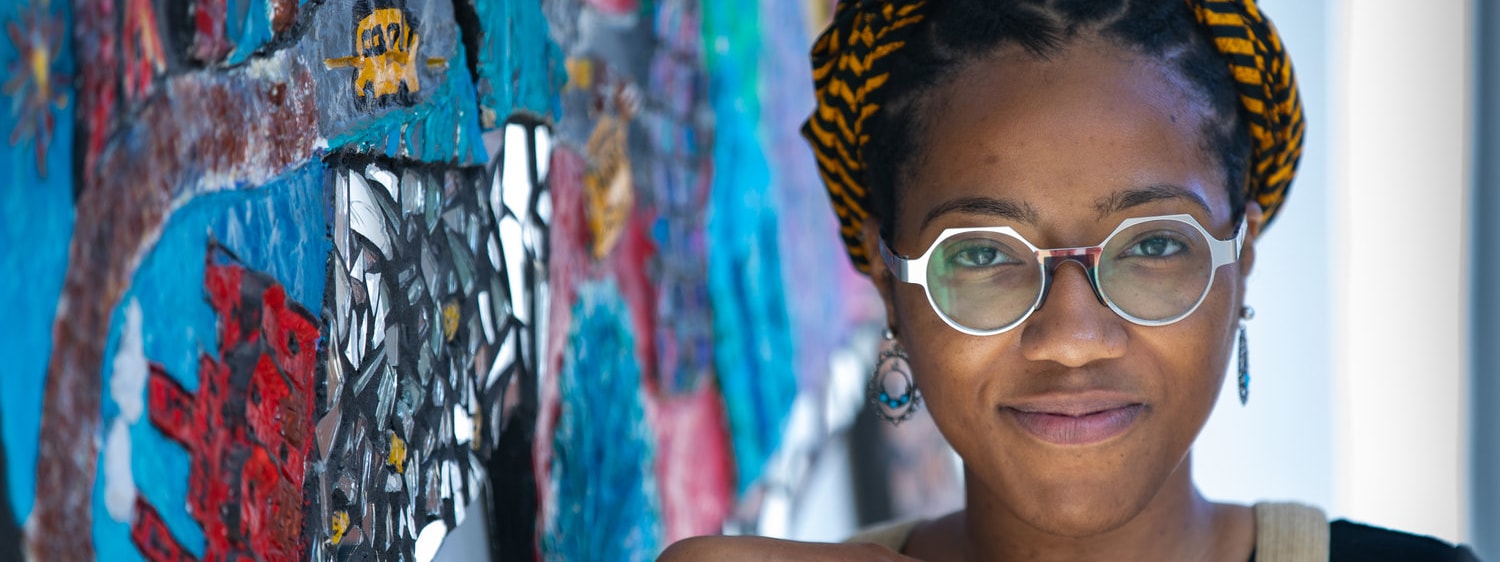 Photo of a Black woman with silver glasses and a multicolored head wrap posing in front of a mural. 