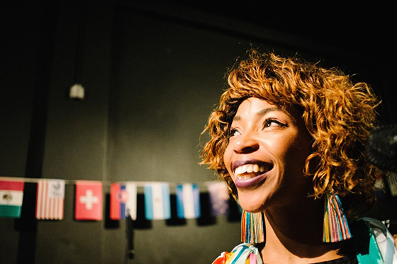 Photo of a Black woman with curly hair and multicolored earrings smiling in front of a wall decorated with international flags.