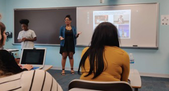 Photo of a woman standing in front of a classroom teaching on a projector.