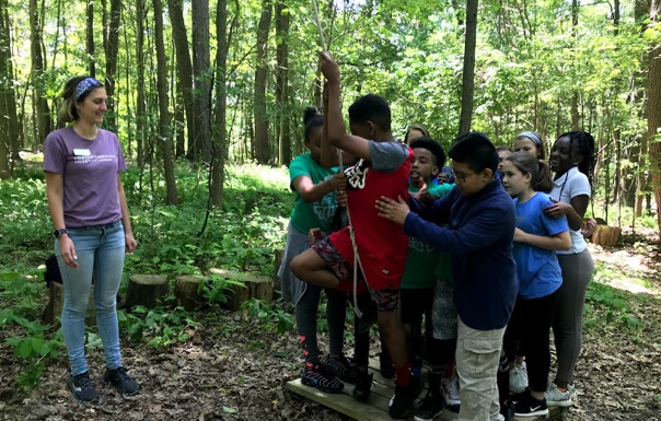 Photo of Chatham University's Lexi Kapla wearing a purple t-shirt while standing in the woods with children who are doing a team-building exercise. 