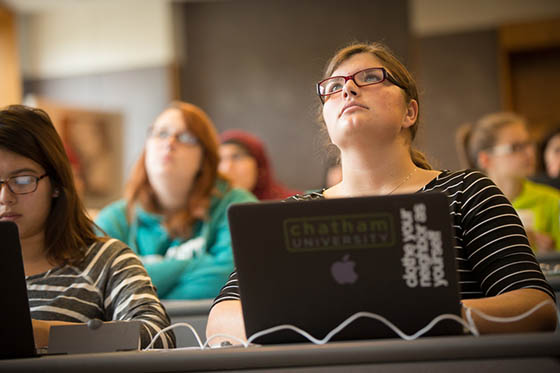 Photo of a Chatham University student paying attention in a lecture hall, with a laptop with a Chatham University sticker on it in front of her