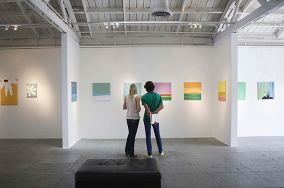 Photo of two young people standing in an art exhibit looking at colorful paintings on the white walls. 