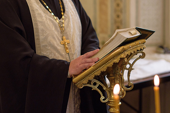 Closeup photo of a priest reading from a bible on a gold stand while wearing a gold cross