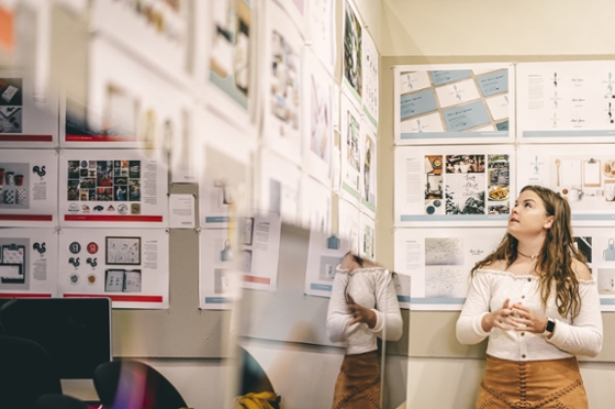 Photo of a Chatham University female student looking up at printed out design projects hung on the wall. 