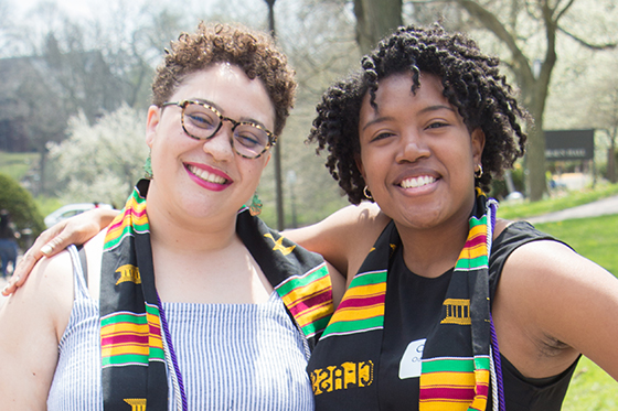 Photo of two Black women, graduating Chatham University students, wearing the multicultural graduation stole and smiling for the camera