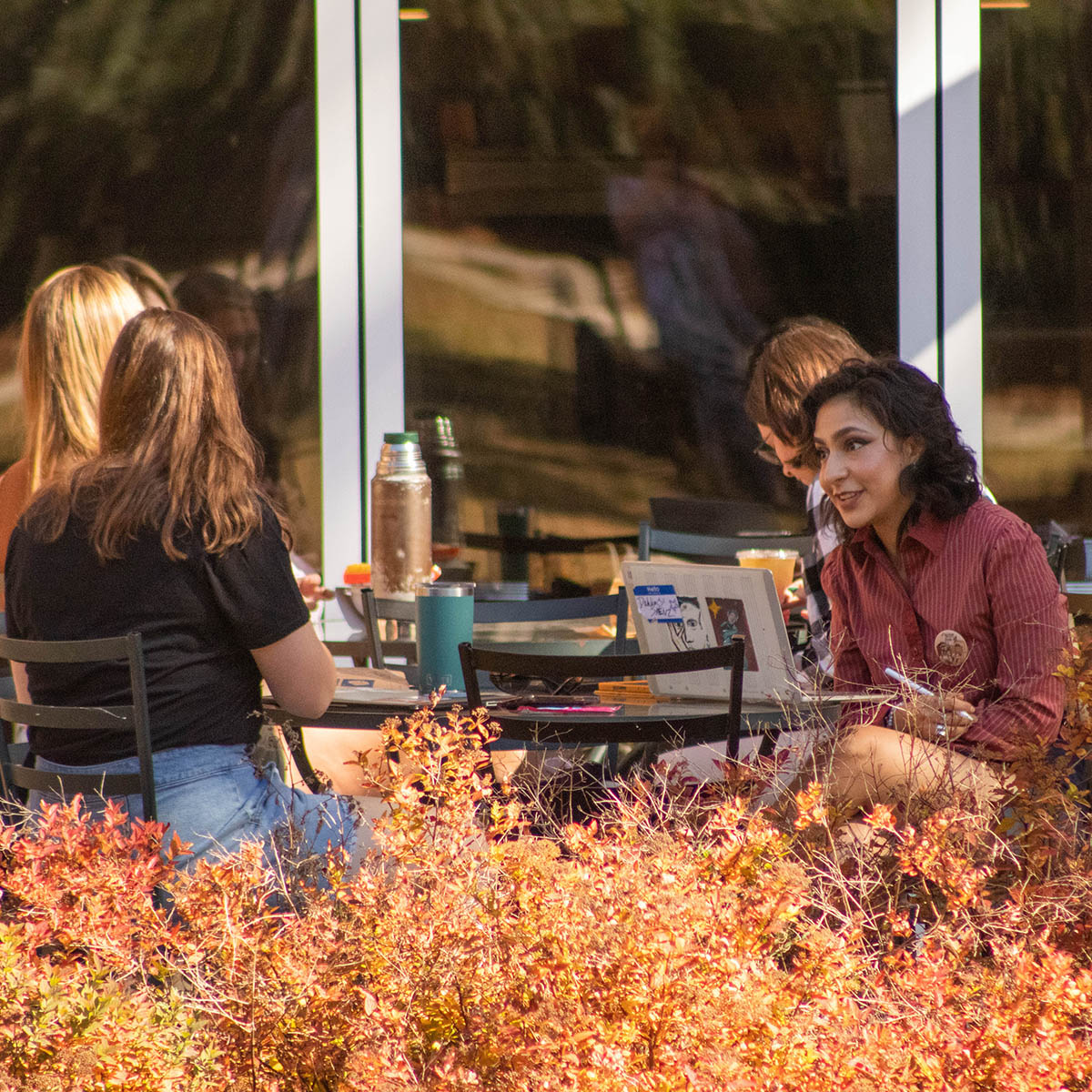 Photo of a group of a students seated outside of Cafe Rachel at tables and chairs, with autumn foliage in the foreground