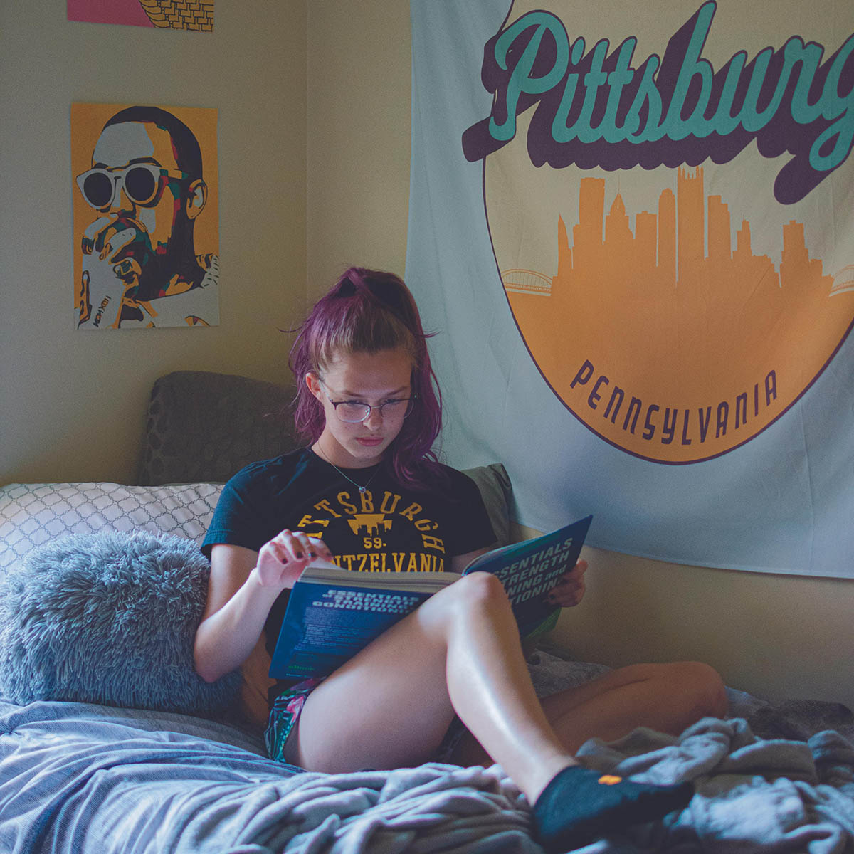 Photo of a young female student with a pink ponytail and glasses reading on her dorm room bed. A large tapestry that says Pittsburgh, Pennsylvania is on the wall