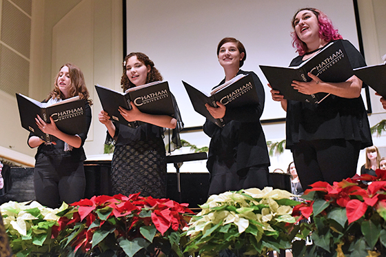 Photo of four Chatham University students singing on stage in a choir while holding music binders and standing in front of an orchestra
