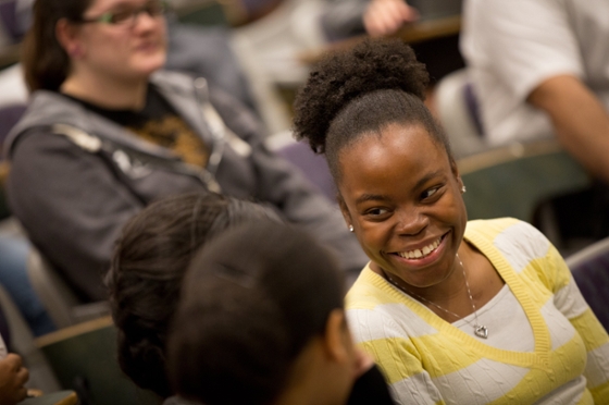 Photo of a woman wearing a yellow sweater, smiling to a friend in a Chatham University lecture hall. 