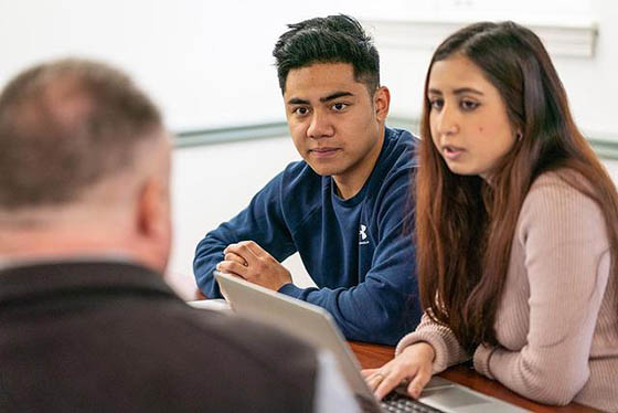 Students and a professor are seated around a table, speaking together