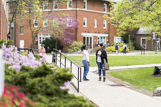 Photo of Chatham University's Shadyside Campus in the spring, with two students speaking to one another beside a red brick academic building