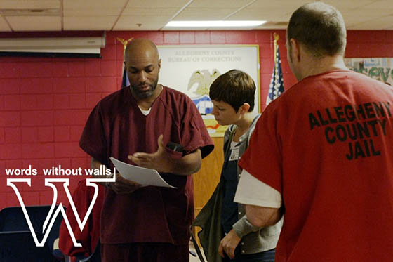 Photo of a Chatham University student speaking to two inmates of Allegheny County Jail, looking at a piece of paper he is holding. Words Without Walls is written on the photo.