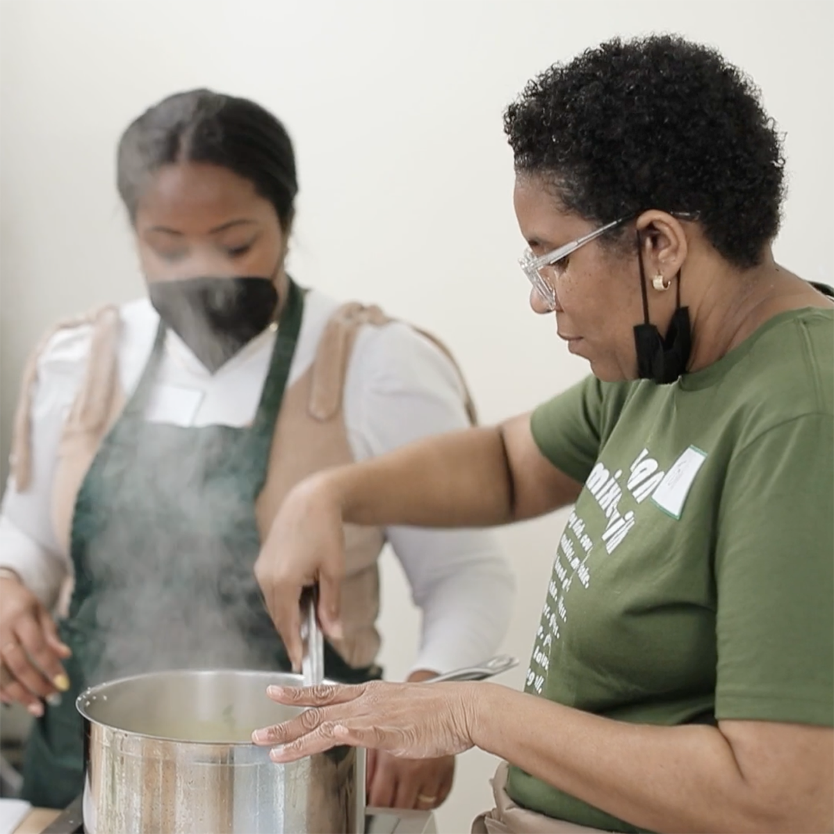 Photo of a Black woman, Toni Simpson, stirring food in a pot and demonstrating to a younger Black woman in an apron and mask who is observing