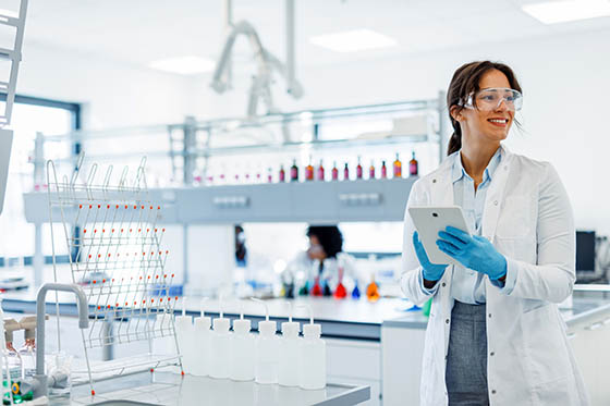 A scientist in a lab coat and gloves uses a digital tablet, smiling as colleagues work in the background.