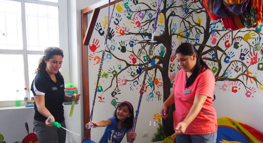 Photo of two women playing with a young girl blowing bubbles in front of colorful mural of hand prints. 