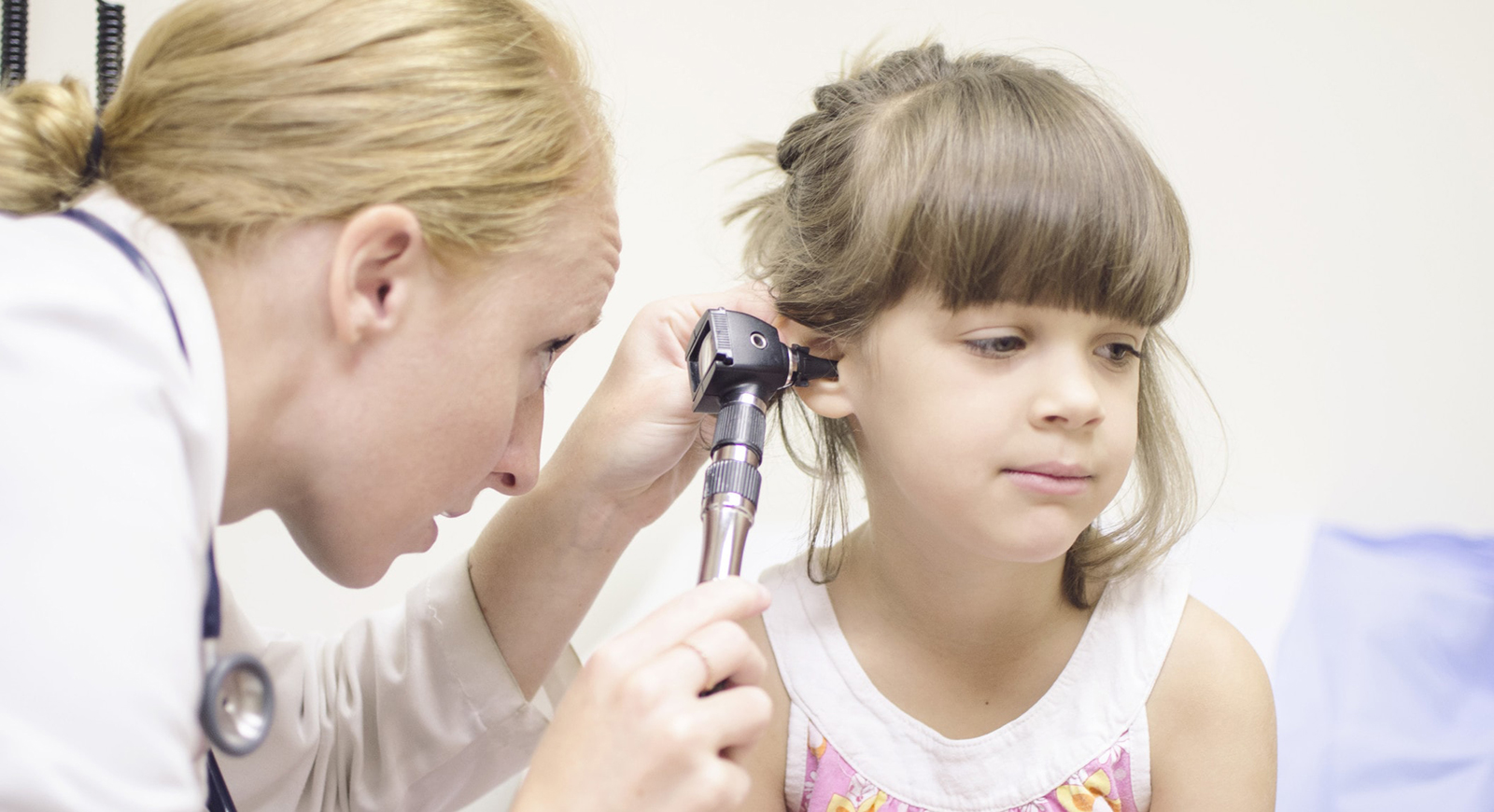 Photo of a medical professional in a white coat looks into a child's ear in an exam room.