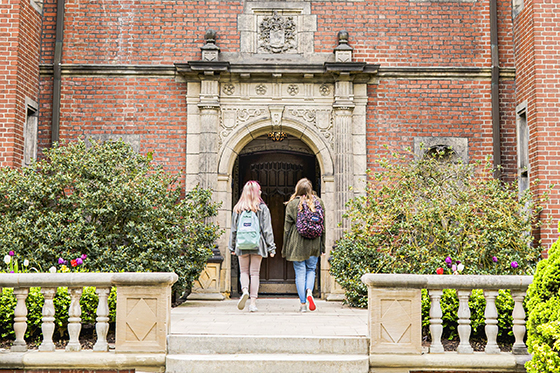 Photo of two female Chatham University students wearing backpacks walking into a red-brick building on Shadyside Campus