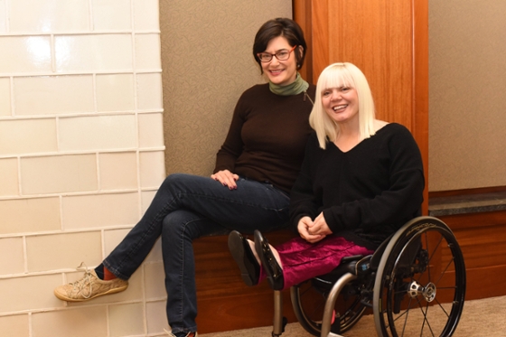 Photo of a woman in a wheelchair and a woman sitting on the arm of it, smiling for the camera