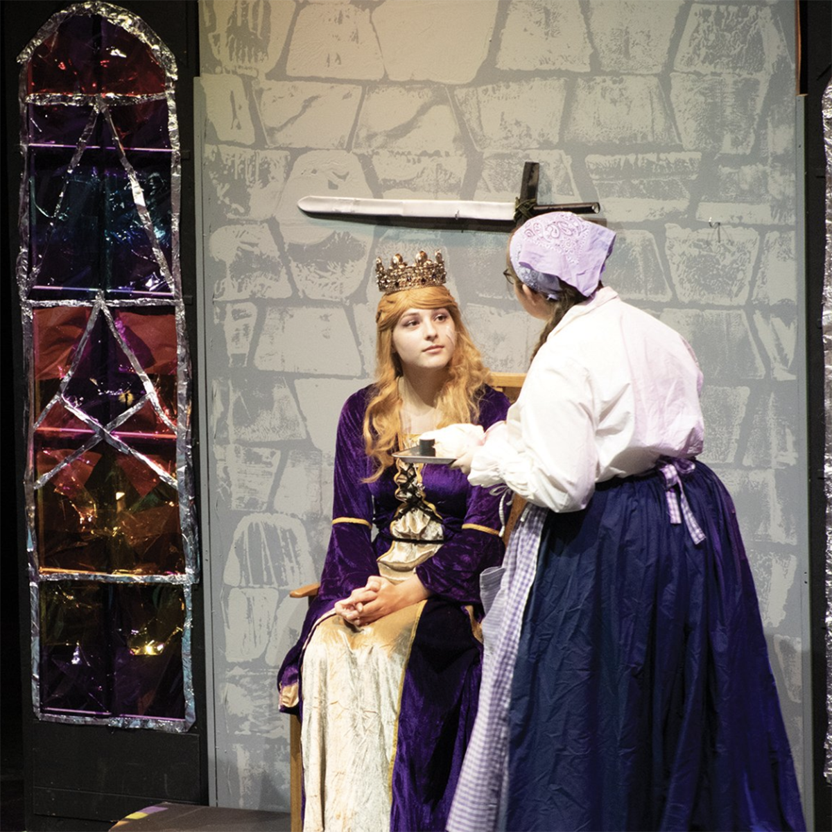 Students perform a play on stage in costume, one wearing a crown and princess gown and the other a skirt and blouse and apron