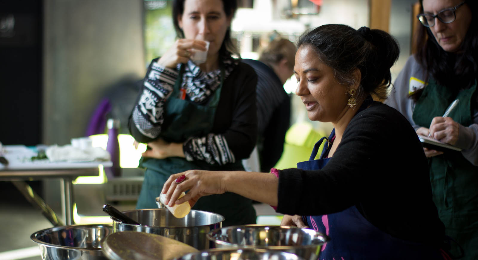 Photo of an Indian woman leading a cooking class, while two women in the background pay attention.
