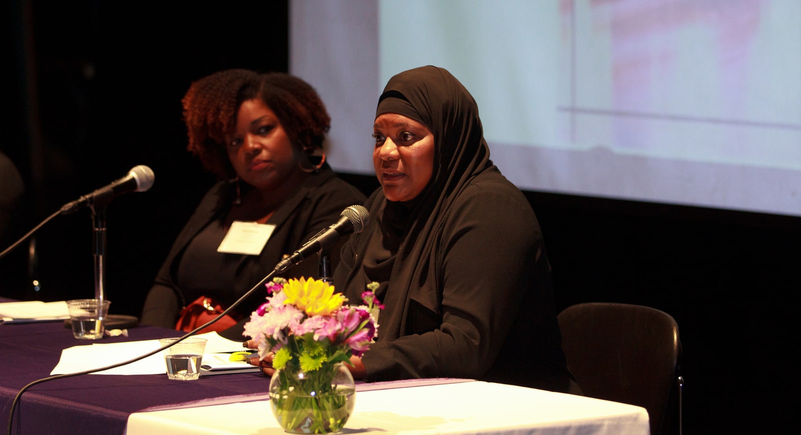 Photo of two Black women on a panel. The hijabi woman is speaking, while the woman beside her listens.