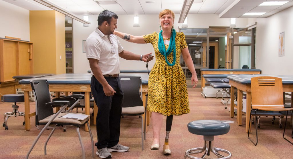 Photo of a Chatham University physical therapy student helping woman with a prosthetic leg walk across an exercise room at Chatham Eastside.