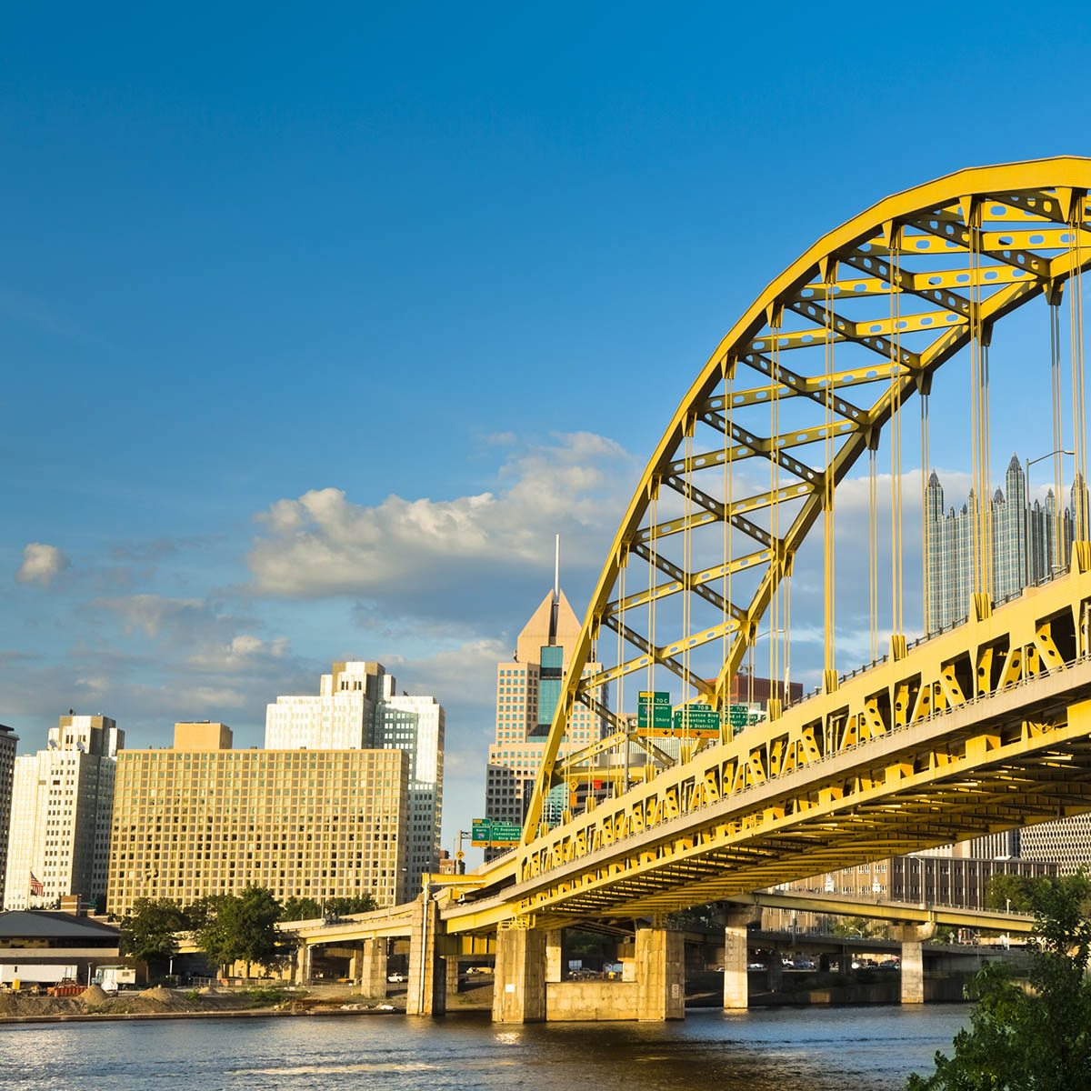 Photo of a yellow bridge over a river and the Pittsburgh skyline