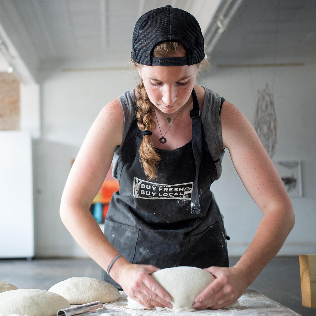 Photo of a young white woman with a black hat, worn backwards, and a long braid is kneading dough on a table