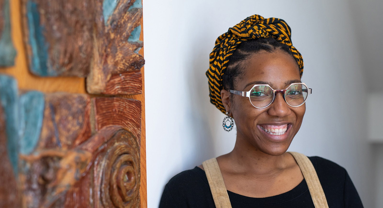 Headshot of Chatham University student Ciera Marie next to a hanging piece of artwork wearing a headscarf and glasses.