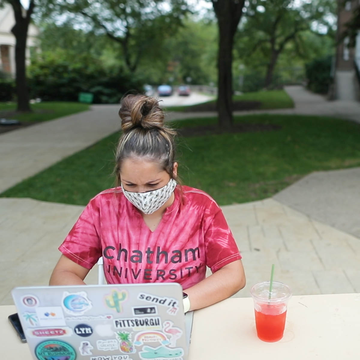 Photo of a young female student in a tie dye Chatham University shirt, wearing a mask and seated at a table in the quad on Shadyside Campus
