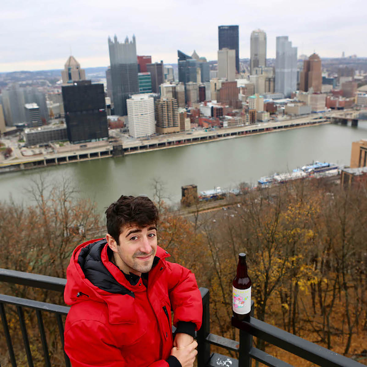 Photo of a young man in a red jacket posing with a beer bottle in front of the Pittsburgh skyline