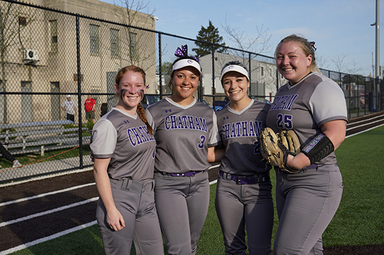 Photo of four Chatham University female softball players pose together in their gray and purple uniforms before a game