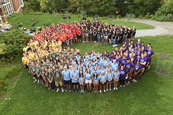 Photo from above, showing a large group of Chatham University students in multicolored T-shirts stand in a C shape together outside on green grass. 
